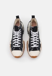 Zapatillas altas de lona negra con cordones blancos, punteras color crema y suela de goma marrón texturizada con picos pronunciados.