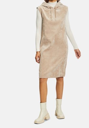 Femme portant une robe sans manches beige à capuche sur un haut à manches longues crème, associée à des bottines crème à semelles épaisses.