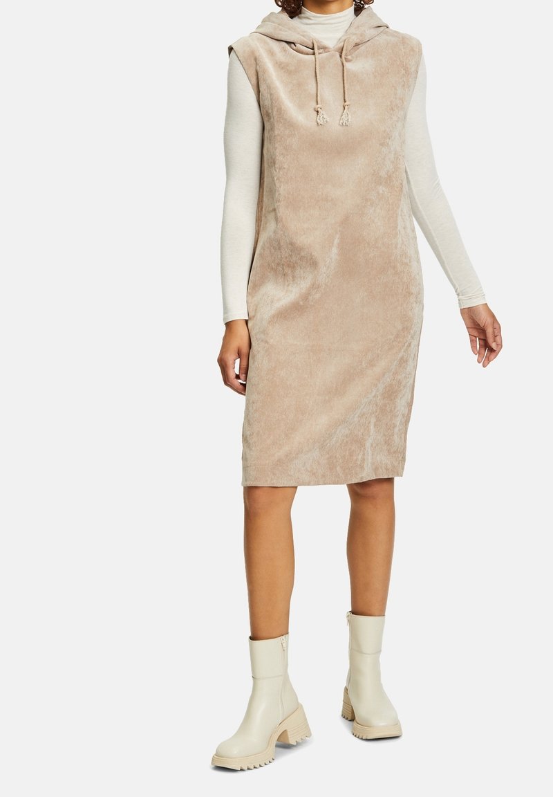 Kvinna iklädd en beige ärmlös klänning med huva över en långärmad krämfärgad topp, kombinerat med krämfärgade ankelstövlar med kraftiga sulor.