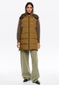 Mujer con chaleco acolchado sin mangas de color marrón sobre un suéter marrón de felpa, pantalones anchos de color oliva y zapatos de punta negra, de pie contra un fondo blanco.