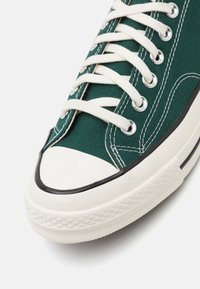 Zelené plátěné tenisky s bílým gumovým špičkovým krytem a mezistélkou, zdobené černým akcentním prošíváním a smetanovými tkaničkami protaženými skrze kovové očko.