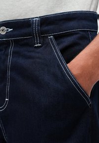 Tmavomodré jeany s bielym šitím, ktoré majú prednú kapsu a zapínanie na gombík v oblasti pása. Textúra sa zdá byť hladká a pevná.