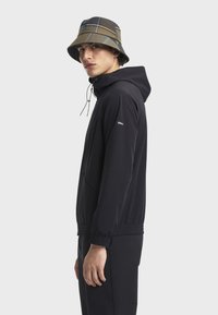 Crna softshell jakna s kapuljačom, elastičnim manžetama i zip džepovima. U kombinaciji s smeškim kockastim šeširom s diskretnim logotipom.