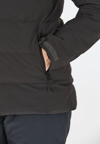 Veste matelassée noire avec une poche zippée et des accents en velcro. Tissu lisse, texture légèrement brillante, avec des coutures ton sur ton et des poignets élastiques.