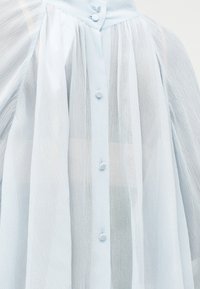 Ανοιχτό μπλε διάφανο μπλουζάκι με υφή υφάσματος, στρογγυλά κουμπιά καλυμμένα με ύφασμα και διακριτικές πιέτες κοντά στον γιακά και τους ώμους.