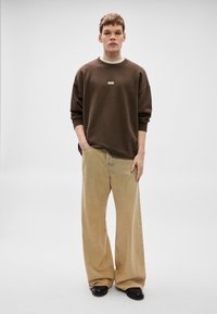 Felpa marrone con una toppa con logo bianco, abbinata a pantaloni larghi beige. La modella è in piedi su uno sfondo neutro, con le mani in tasca.