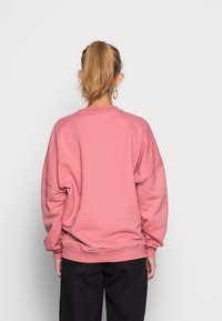Sudadera rosa de gran tamaño con una textura suave, hombros caídos y puños acanalados. Combinada con pantalones oscuros, mostrando un ajuste relajado.
