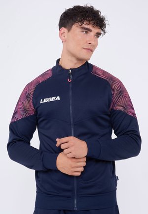 Giovane uomo con capelli corti e ricci che indossa una giacca blu navy con zip con motivi geometrici rosa sulle spalle e il logo "LEGEA" sul petto.