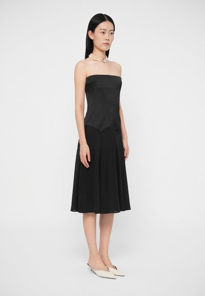 Žena stojaca v čiernych midi šatách bez ramienok, s vrstveným náhrdelníkom z perál a bielymi bodkovanými lodičkami na jednoduchom pozadí.