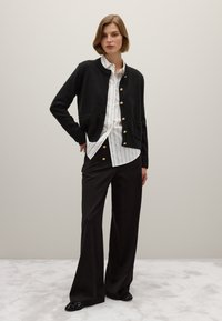 Cardigan noir en tricot avec col rond et boutons dorés, associé à une chemise rayée blanche et un pantalon large noir. Détails de tissu texturé.
