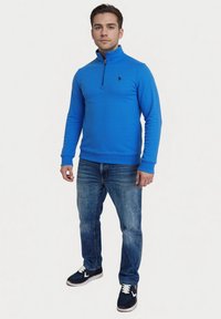 Blauwe, ritssluiting pullover gemaakt van zacht materiaal, gecombineerd met blauwe jeans en donkere sneakers. Heeft een klein zwart logo op de borst.