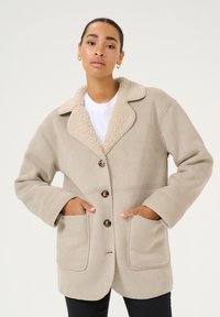 Beige takki, jossa on pörröinen kermahvärinen kaulus, neljä nappia, kaksi etutaskua ja pehmeä, teksturoidun pinnan viimeistely.