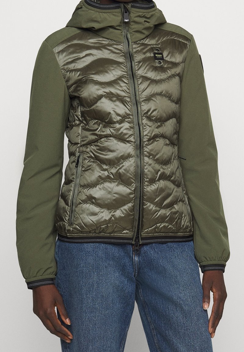 Gevoerde jas met een gewatteerde olijfgroene voorkant, gladde mouwen en een capuchon. Voorzien van een ritssluiting aan de voorkant, zijzakken en elastische boorden.
