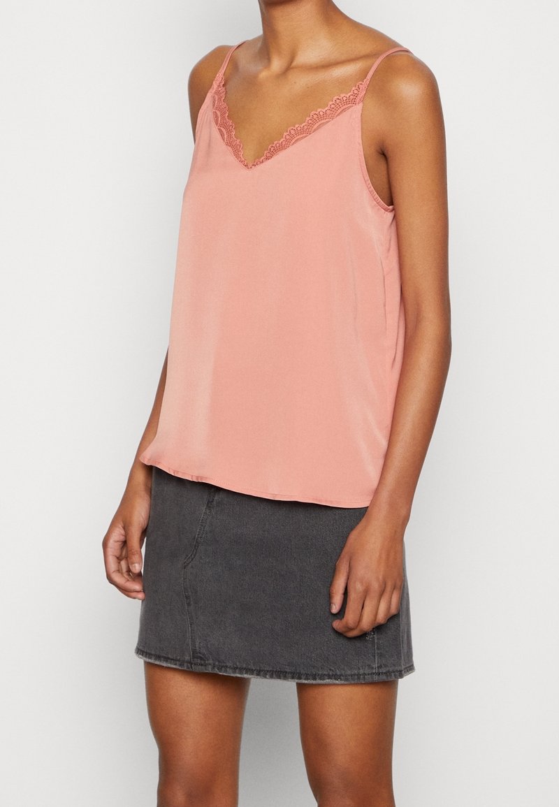 Femme portant un caraco rose sans manches avec bordure en dentelle, associé à une mini-jupe en jean noire, debout devant un fond uni.