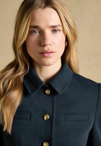 Blonde woman with blue eyes wearing dark navy jacket with gold buttons, posing against a beige background.