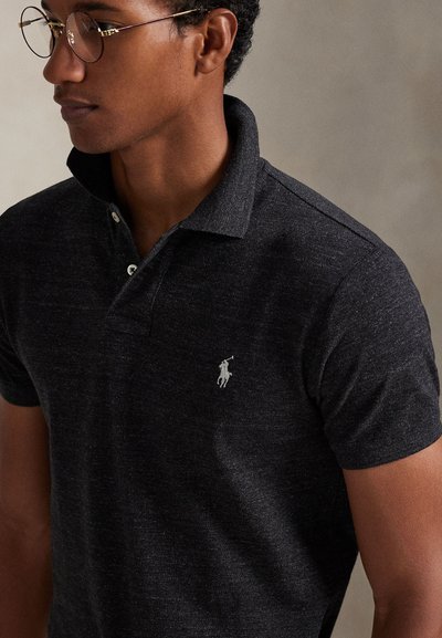 Young man wearing round glasses and a dark gray polo shirt with a small white embroidered logo on the chest.