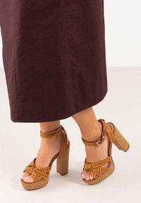 Sandalias de plataforma trenzadas en marrón con un grueso tacón cuadrado, que presentan un diseño trenzado y una correa en el tobillo con detalle de hebilla.