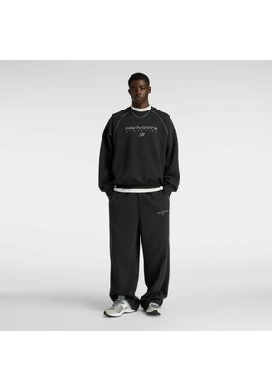 Junger Mann steht und trägt ein schwarzes New Balance Sweatshirt und dazu passende weite schwarze Hosen mit weißen und grauen Turnschuhen vor einem einfarbigen Hintergrund.