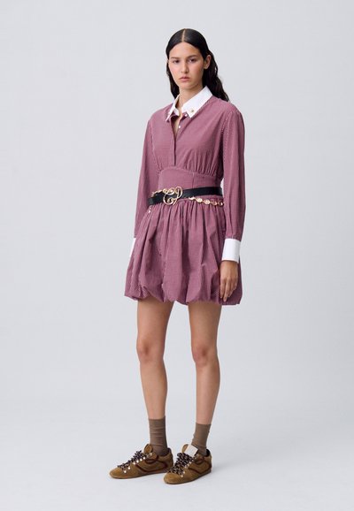 Modella in un vestito a quadretti rosso e bianco, corto, con vita stretta e cintura nera, abbinato a scarpe da ginnastica marroni e calzini grigi.