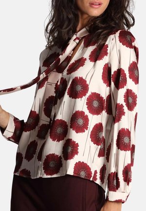 Blusa a maniche lunghe in tessuto crema con motivo floreale rosso, con scollo a fiocco e vestibilità ampia, polsini arricciati e texture morbida.