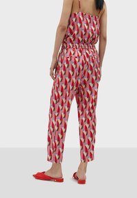 Kvinna iförd byxor och topp med röd geometrisk mönstring, parat med röda slip-on platta skor, stående mot en enkel bakgrund.