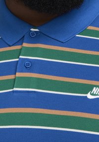 Gros plan sur un polo à rayures bleues, vertes et beiges avec des boutons bleus et un logo Nike partiellement visible sur la poitrine.