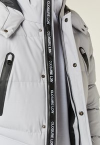 Light grey insulated jacket featuring a hood, black zipper, and logo detailing on the front opening. Smooth texture with functional pockets.
