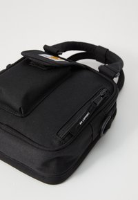 Sac rectangulaire noir avec une poignée supérieure, un rabat avant et une fermeture éclair sur le côté. Fabriqué en tissu texturé avec une étiquette et des accents lisses.