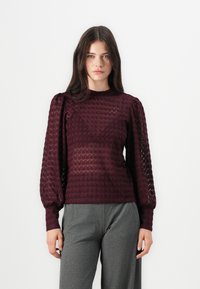 Bordowy sweter o fakturowanym splocie z bufiastymi rękawami, z geometrycznym wzorem i dopasowanym krojem. W zestawie z szerokimi spodniami w kolorze szarym.