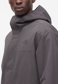 Homme portant une veste à capuche grise North Face avec le logo visible sur la poitrine, montré des épaules vers le haut.