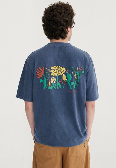 Homme aux cheveux bouclés portant un t-shirt bleu avec des motifs floraux colorés dans le dos, associé à un pantalon marron, debout de dos.
