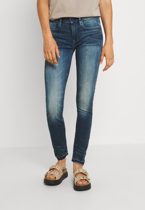 Calças skinny em denim azul desbotado com ligeiras desgastes, apresentando um design clássico de cinco bolsos e uma cintura de altura média. Combinadas com sandálias de cor clara.