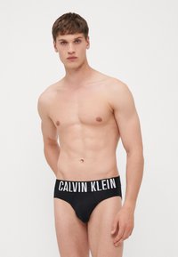 Schwarze Baumwollslips mit weißem "CALVIN KLEIN"-Logo am Bund, enganliegendes Design, glatte Textur und niedriger Schnitt. Modell steht vor einem weißen Hintergrund.