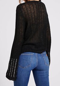 Pull en tricot noir avec une coupe ample, texture côtelée et manches évasées ornées de sequins. Associé à un jean en denim bleu.