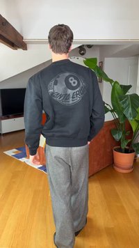 Vīrietis, kurš valkā melnu Santa Cruz džemperi un pelēkas bikses, staigā dzīvojamā istabā ar koka grīdu, oranžu dīvānu un lielu zaļu augu.