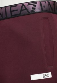 Burgundske atletske kratke hlače izrađene od rastezljivog materijala. Imaju crni elastični pojas s logom i bijelu oznaku s logom sprijeda.