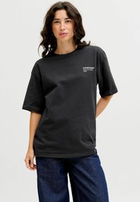 Sort bomulds T-shirt med løs pasform, korte ærmer, rund halsudskæring og en broderet logo foran i hvid. Sammen med blå denim bukser.