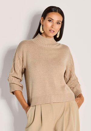 Frau mit langen dunklen Haaren, die einen beigen Strickpullover mit Kabelärmeln, beigen plissierten Hosen und großen goldenen Tropfenohrringen trägt.