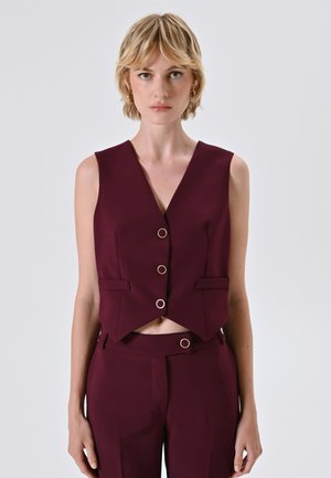 Pullover bordeaux a vita alta con scollatura a V, caratterizzato da dettagli con bottoni dorati e tasche laterali. Realizzato in un tessuto liscio e strutturato.