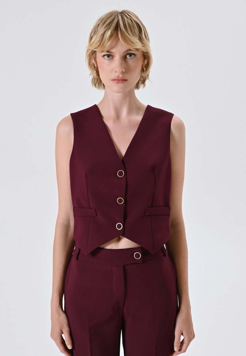 Pullover bordeaux a vita alta con scollatura a V, caratterizzato da dettagli con bottoni dorati e tasche laterali. Realizzato in un tessuto liscio e strutturato.