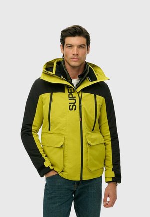 Geel en zwarte hooded jas met twee voorzakken, een ritssluiting en het woord "SUPER" prominent op de borst gedrukt.