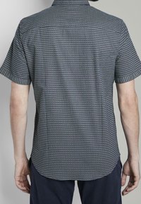 Camicia a maniche corte di colore blu navy con un motivo geometrico in bianco e blu chiaro. Tessuto morbido con colletto classico e un orlo leggermente curvato.
