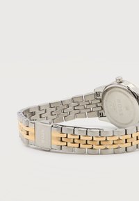 Μεταλλικό μπρασελέ ρολογιού σε ασημί και χρυσό με κούμπωμα χαραγμένο με τη λέξη «BOSS», προσαρτημένο στην πίσω πλευρά μιας ασημί θήκης ρολογιού που βρίσκεται ανάποδα.