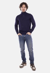 Maglione a zip blu navy, design aderente, polsini e orlo a coste, abbinato a jeans azzurri e scarpe da ginnastica marroni.