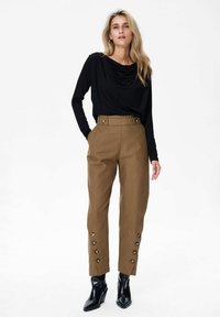 Zwarte lange mouw top met een gedrapeerde halslijn, gecombineerd met bruine hoge taille broek met gouden knoopdetails aan de zijkant en zwarte enkellaarsjes.