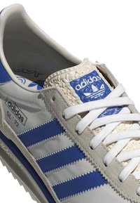 Tenisky Adidas SL-72 mají šedou základnu, modré pruhy, texturované bílé detaily a jazyk s vytlačeným logem, doplněný bílými tkaničkami.