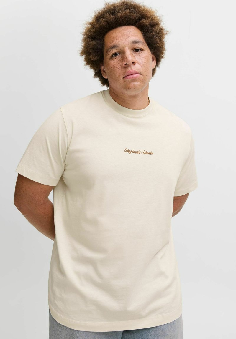 T-shirt en coton couleur crème avec des manches courtes et un col rond, arborant un petit logo en texte marron "Originals Studios" sur la poitrine.