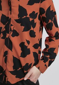 Camicia a maniche lunghe in ruggine con finitura testurizzata, caratterizzata da grandi motivi floreali neri. Chiusura con bottoni visibile vicino al collo.