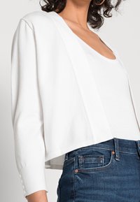 Biała, krótkia kardiganka o ribbowanej strukturze i długich rękawach, z zapięciami na guzikach przy mankietach. Połączona z prostą białą bluzką i ciemnymi dżinsami.