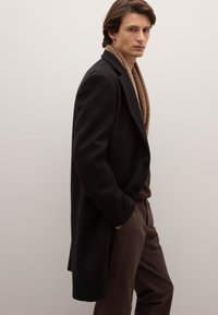 Abrigo largo de lana negra con solapas de muesca, bolsillos laterales y cierre de un solo botón, llevado sobre un jersey de cuello alto marrón texturizado y pantalones a juego.